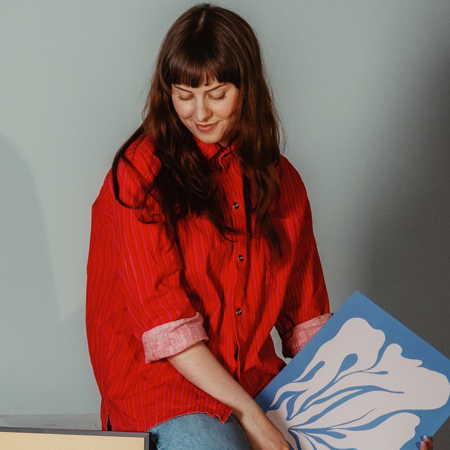 Woman in a red shirt holding a blue and white abstract painting against a gray background