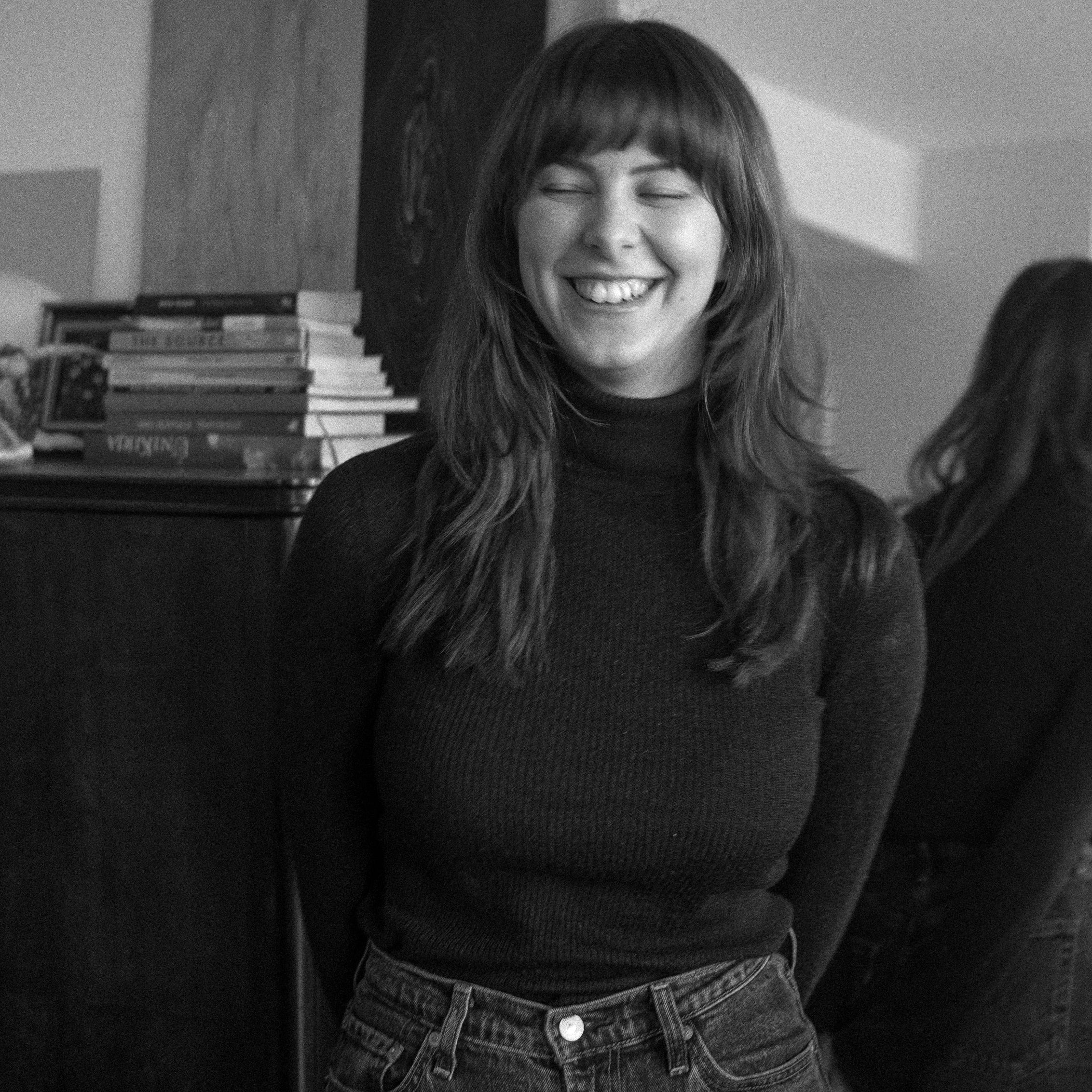 Black and white photo of a woman smiling in an indoor setting with books and a painting in the background.